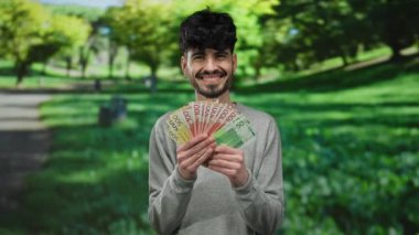 Norveç kronu banknotları tutarken gülümseyen bir adam yemyeşil bir park ortamında, Norveç 'te açık havada mutluluk ve finansal başarı sergiliyor..