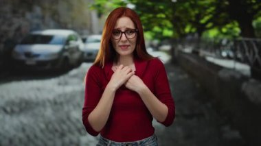 Kızıl saçlı, gözlüklü ve kırmızı kazaklı bir kadın güneşli bir günde arka planda arabalarla şehir caddesinde duruyor..