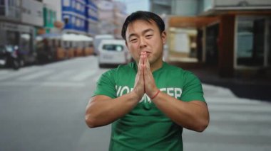 Yeşil gönüllü gömleği giyen genç adam şehir sokağında duruyor ve arkasında renkli binalar olan bir dua jesti gösteriyor..