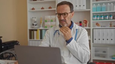 İspanyol erkek doktor, tıbbi ekipman ve kitaplarla çevrili bir dizüstü bilgisayar kullanırken beyaz önlük ve gözlük takarak bir hastane odasına odaklanmış oturuyor..