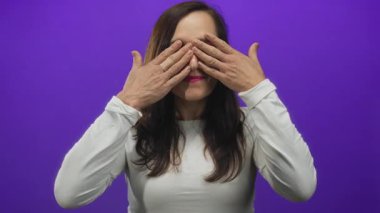 İspanyol kadın bir stüdyo ortamında mor bir duvara karşı iki eliyle gözlerini kapatır; sürpriz şaşkınlık merak.