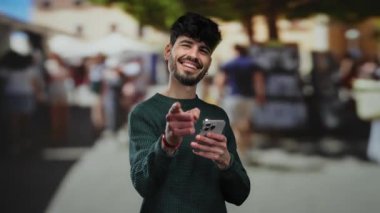 Şehir sokağındaki kameraya gülümseyen genç adam akıllı telefon tutarak canlı şehir manzarasını resmediyor