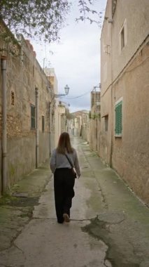 Mallorca 'da taştan binalarla kaplı, büyüleyici bir sokakta yürüyen bir kadın hafif bulutlu bir gökyüzünün altında huzurlu bir Akdeniz atmosferi yayıyor..