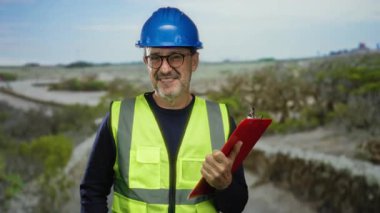 Açık havada, yüksek görüş açısına sahip yelek giyen ve elinde pano olan bir adam, doğal bir manzara arkaplanına gülümseyen, kırsal alanda inşaat ve güvenlik ekipmanlarını gösteren bir adam..