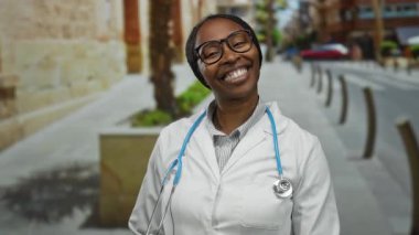 Şehrin arka planında steteskop ve gözlük takarak güvenle gülümseyen kadın doktor. Tarihi mimari ve ağaçlar sağlık merkezini yakalıyor.