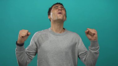 Neşeli ve kendine güvenen genç bir adam canlı deniz mavisi arkaplanına karşı yumruklarını sıkarak başarı ve coşkuyla iletişim kuruyor..