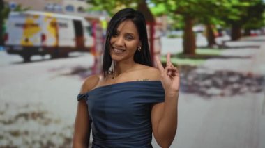 Gülümseyen ve barış işareti yapan bir kadın, güneşli bir günde arka planda ağaçlar ve renkli bir minibüsle mutluluk ve güven saçan bir şehir caddesinde..