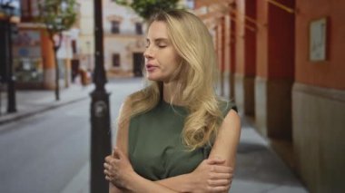 Sarı saçlı bir kadın şehir caddesinde gergin görünüyor. Açık havada kollarını kavuşturuyor. Sıradan kıyafetleri ve ilgi çekici bir çevresi olan tipik bir şehir sahnesini vurguluyor..