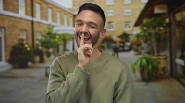 Genç adam neşeli bir şekilde gülümsüyor. Canlı bir şehir caddesinde parmaklarıyla. Açık havada genç ve samimi bir enerji ve şehir yaşamını yakalıyor..