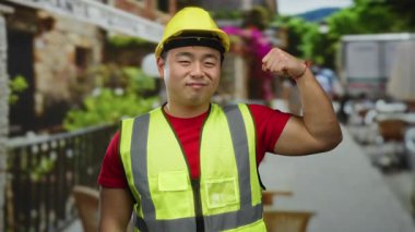 İnşaat malzemesi giymiş bir adam sokak ortasında kaslarını esnetiyor, sarı bir şapka ve yansıtıcı yelek giyerken gücünü ve güvenini gösteriyor..