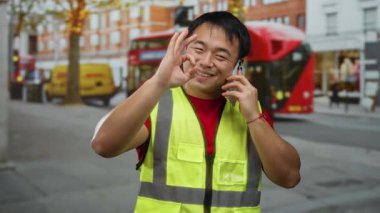 Güvenli yelek giyen bir adam akıllı telefonuyla telefon ederken gülümsüyor ve el kol hareketi yapıyor. Meşgul şehir caddesindeki sembolik kırmızı çift katlı otobüs ve teslimat minibüsleri görünür durumda..