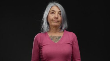 Yaşlı, gri saçlı, dövmeli ve pembe kazaklı bir kadın. Stüdyo ortamında derin düşüncelere daldığını ifade eden siyah bir arkaplan karşısında izole edilmiş..