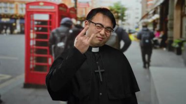 Arka planda ikonik kırmızı telefon kulübesi olan işlek bir caddede güvenle poz veren bir rahip.