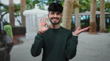 Sakallı genç adam gülümsüyor. Güneşli bir şehir caddesinde dikiliyor. Arka planda ağaçlar var..