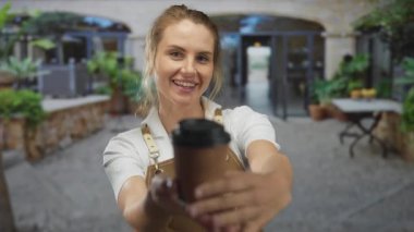 Açık havada önlükle gülümseyen bir kadın güneşli bir şehir sokağında yeşil ve taş binalarla çevrili masaları olan bir kahve fincanı sunuyor..