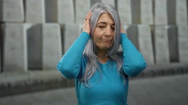 Dışarıdaki kadın, modern şehir arka planına karşı sakin bir ifadeyle kulaklarını kapatıyor, canlı mavi bir kazak giyiyor, şehir gürültüsünün ortasında sükuneti somutlaştırıyor..