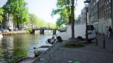 Amsterdam 'daki bulanık şehir manzarası beyaz bir minibüs ve ilkbaharda caddeyi kaplayan ağaçlarla çevrili bir kanal gösteriyor..