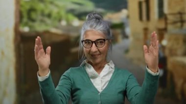 Gri saçlı, gözlüklü yaşlı bir kadın canlı bir sokakta jestler yapıyor. Arka planda bulanık binalar ve yemyeşil alanlar var. Açık hava kentsel ortamı öneriyor..