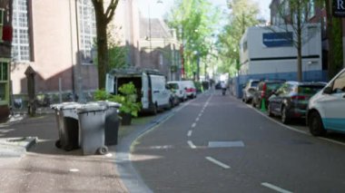 Ağaçlar ve binalar tarafından çerçevelenmiş, park halindeki arabalar ve çöp kutularıyla dolu boş bir Avrupa şehir caddesinin bulanık görüntüsü, kentsel atmosferi dağıtıyor..