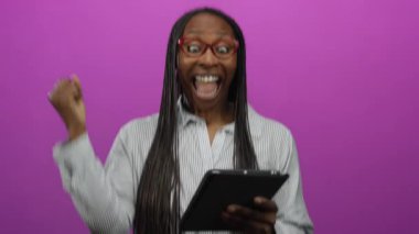Başarılı bir kadının pembe arka planda tableti tutması, heyecan verici bir şekilde yumruk sallaması, neşeli bir gülümseme, duygu ve mutluluk göstermesi..