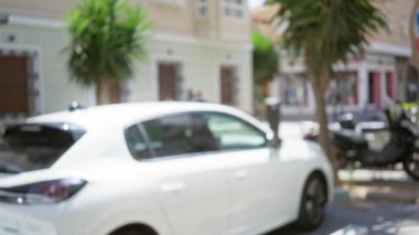 Arka planda, palmiye ağaçları ve binalarla dolu bir caddeye park edilmiş bulanık beyaz bir araba odak dışı ve bokeh efektli kentsel manzara