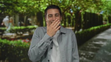 Parkta arka planda ağaçlarla gülümseyen, neşeli bir ortamda mutluluk ve doğa taşıyan genç İspanyol bir adam.