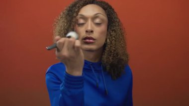 Makyaj fırçasını turuncu stüdyoda yüzüne kıvırcık saç çerçeveli yumuşak ışıklandırma altında yanak yanağa süren kadın: kendine güven ve özgüven.