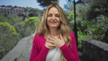 Parkta mutlu bir şekilde gülümseyen bir kadın güneşli bir günde pembe bir kazak giyiyor. Etrafında canlı yeşillikler ve ağaçlar var..