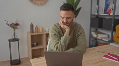 Genç İspanyol bir adam rahat bir oturma odasında esniyor. Ev dekoru ve renkli defterlerle çevrili bir dizüstü bilgisayarla ahşap bir masada otururken yorgun görünüyor..