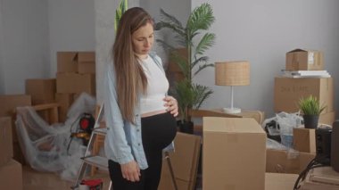 Hamile kadın yeni evin oturma odasında kutuları düzenliyor, paketlenmemiş eşyalarla çevrili, parlak iç mekanda beklenti ve ev yapımı..
