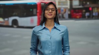 Mavi tişörtlü, gözlüklü, kalp işareti yapan arka planda bulanık otobüsle gezen genç bir kadın canlı bir şehir manzarası sergiliyor..