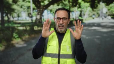 Yansıtıcı yelek giyen gözlüklü adam, ağaçlar ve gölgelerle dolu bir parkta dikilirken ürkmüş görünüyor..