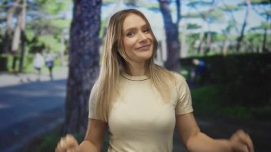 Gündelik kıyafetli genç sarışın kadın güneşli bir parkta ağaçlarla ve bulanık figürlerle dikilirken güzel bir günde arka planda yürürken tırnak işareti yapıyor..
