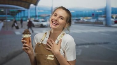 Havaalanının dışında elinde dondurma külahıyla gülümseyen bir kadın arka planda gezginlerin olduğu hareketli bir ulaşım merkezinde neşe ve rahatlık gösteriyor..