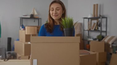 Mavi elbiseli kadın, yeni apartman dairesinde taşınan kutuları taşıyor. Etrafı tıka basa doldurulmuş eşyalarla çevrili ve elinde bitki tutarken gülümsüyor..