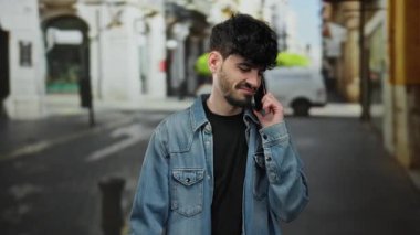 Sakallı genç adam kot ceket giyiyor. Şehir merkezinde telefonla konuşuyor. Arka planı bulanık..