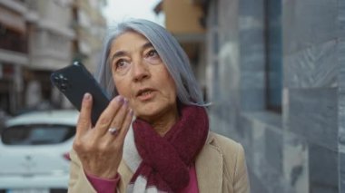 Gri saçlı yaşlı bir kadın, şehir caddesinde atkı ve palto giyip, iletişim ve şehir hayatını anlatırken akıllı telefona konuşur..