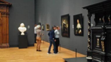 Müzenin içindeki bulanık görüntü, loş aydınlatılmış bir galeride sanat eserleri keşfeden, yumuşak bokeh ışıklandırması altında çözülmüş resimler ve heykeller sergileyen insanların görüntüsü..