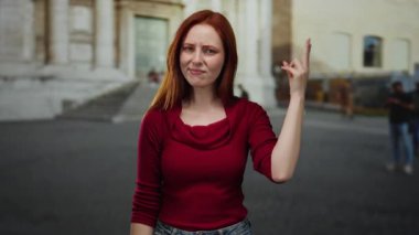 Şehir sokaklarında dikilen kızıl saçlı ve kırmızı kazaklı kadın, mimari ve uzak yayalarla çevrili şehir ortamında poz verirken meraklı görünüyor..