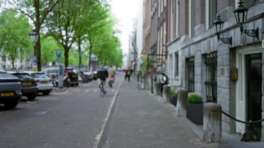 Bulanık yayalar ve bisikletçiler Amsterdam, Hollanda 'da bulutlu bir gökyüzü altında klasik mimari, park edilmiş arabalar ve yemyeşil sergilenen pitoresk bir caddede.