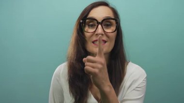 Dudakları üzerinde parmaklarıyla sessizlik gösteren, gözlüklü, izole edilmiş mavi duvar arkaplanı olan bir kadın..