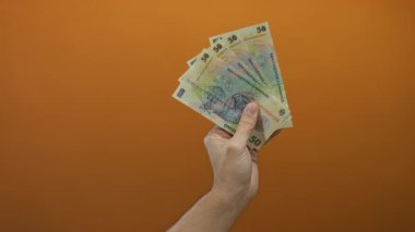 Kafkasyalı bir adam, Romen lei banknotlarını izole edilmiş turuncu bir arka plana karşı tutuyor. Finansal konsept ve para birimini canlı bir iç mekan ortamında sergiliyor..