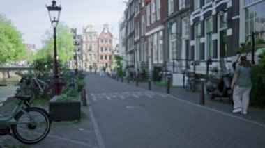 Amsterdam 'ın bulanık şehir manzarası. Kanal boyunca yürüyen insanlar, bisiklet vitrinleri, klasik mimari ve ilkbaharda huzurlu bir şehir havası..