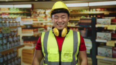 Güvenlik teçhizatı olan genç bir adam markette gülümsüyor ve arka planda raflar dolusu yiyecek var. Bu da içeride inşaat veya bakım işi olduğunu gösteriyor.