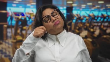 Beyaz tişörtlü bir kadın spor salonunda üzgün üzgün bir ifade sergiliyor. Etrafı bulanık egzersiz ekipmanlarıyla çevrili. Kapalı alanda duygusal bir an yakalıyor..