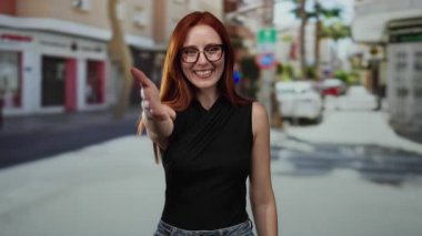 Kızıl saçlı ve gözlüklü bir kadın şehir caddesinde sıcacık gülümsüyor, günlük kıyafetler giyiyor, dostça bir el uzatıyor, şehir dışı cazibesini ve sosyal ambiyansı sergiliyor..