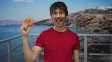 Kırmızı tişörtlü, gülümseyen genç adam, iskelede tırabzan yaparak jöleli çöreği tutuyor. Mutluluk..