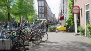 Amsterdam 'ın güneşli bir caddesinde park halindeki bisikletlerin yanından geçen insanların bulanık görüntüsü kaldırımda ağaçların oluşturduğu canlı ama sakin şehir atmosferini yakalıyor..