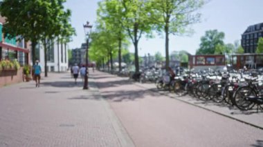 Hollanda, Amsterdam 'da ağaçlık bir cadde boyunca yürüyen insanların bulanık görüntüsü. Arka planda bisikletleri park edilmiş ve tarihi binalar görülebiliyor..