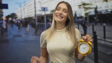 Sarı alarmlı saat tutan kadın şehir sokaklarında omuz silkme hareketi sergiliyor. Şehir çevresi ve günlük koşuşturma arasında kaygısız bir ifadeyi vurguluyor..